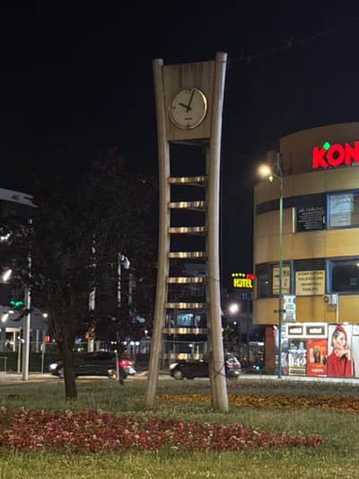 This image captures a street scene at night in Ilidža, Bosnia and Herzegovina. The focal point is a tall, modern clock tower with a light-colored frame and a circular clock face displaying the time as approximately 12:10. To the right of the clock tower is a multi-story yellow building with illuminated signage. One prominent sign reads "KONGRESNI" in large red letters. Below this, other signs are visible, including one that says "HOTEL". The building also has windows displaying advertisements, and an air conditioning unit is visible on the upper facade. In the foreground, there is a well-maintained grassy area with flowerbeds featuring red and yellow foliage. Several cars are parked on the street or driving by. A dark-colored car with its headlights on is visible on the left side of the image, and another car is partially visible on the right. Trees with dark foliage are scattered throughout the scene. The overall atmosphere is that of a quiet urban night, with artificial lighting from the buildings and streetlights illuminating the surroundings.