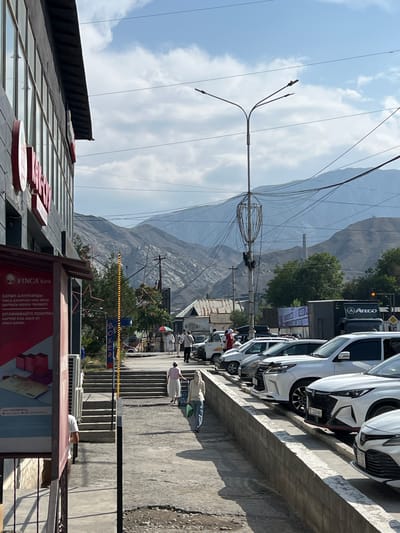 The image captures a street scene in Kadamjay, Kyrgyzstan. On the left side of the frame, a modern building with large windows and prominent signage for "FINCA Bank" is visible. A sidewalk runs alongside the building, leading towards a set of stairs. Several people are walking on the sidewalk, some carrying bags. A variety of cars, primarily white SUVs and sedans, are parked along the street on the right side of the image. In the background, a range of rugged, mountainous terrain is present, with power lines crisscrossing the sky. The weather appears to be fair with scattered clouds. The overall impression is that of a bustling urban area situated against a backdrop of natural landscapes.