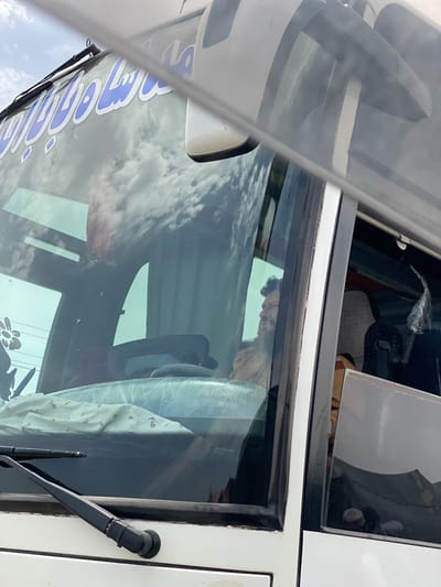 The image captures the front-left exterior of a white bus or large passenger van, viewed from a slightly elevated perspective. The sky, featuring white cumulus clouds, is vividly reflected across the vehicle's large windshield, indicating a bright daytime setting.

Inside the vehicle, a man with a beard is visible, seated in the front, likely in the driver's seat or front passenger seat. He looks towards the right of the frame, appearing to be at rest or waiting. Part of the dashboard is covered by a light-colored, patterned fabric, possibly a blanket or sheet. Black windshield wipers are positioned at the base of the windshield.

A white side mirror is partially visible on the right side of the frame. On the top portion of the windshield, blue text in Arabic or Persian script is displayed, which reads "شاه مسافر بری" (Shah Musafir Bari), meaning "Royal Passenger Transport" or "King's Passenger Transport," commonly used for bus services. The scene depicts a stationary vehicle in an outdoor environment, consistent with the provided location context of Panjwai, Afghanistan.