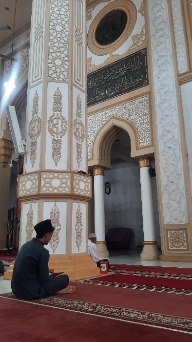 The image captures an indoor scene within what appears to be a mosque or Islamic prayer hall, characterized by ornate Islamic architecture and a person engaged in prayer. The perspective is from slightly behind and to the right of the individual.

In the foreground, a person is seated on a red prayer carpet, facing towards the left side of the frame. They are wearing dark clothing, possibly a tunic or long shirt, and a dark cap (kufi or peci). Their bare feet are visible, and they are in a traditional seated prayer posture, likely *jalsa* (sitting between prostrations) or *tashahhud* (the final sitting position in prayer). The red carpet features intricate gold or yellow patterns along its edges and possibly marking prayer lines.

To the left of the praying individual, a small wooden lectern or stand is visible. Beyond this, partially obscured, is a white garment or fabric draped over something, next to a small red object.

The background and left side of the image showcase the elaborate interior design of the building. Walls and columns are adorned with intricate Islamic geometric patterns and calligraphy, primarily in white, beige, and light brown tones. A prominent feature is a large rectangular panel mounted on a wall or column, displaying Arabic calligraphy in gold or light-colored script against a dark, possibly black, background. To its left, a large oval decorative element, dark in its center with a golden-beige border, is visible.

Further into the background, large, smooth white columns with golden capitals or bases support an archway, suggesting a transition to another part of the building. More geometric patterns, including star-like motifs, are visible on various panels and sections of the architecture. A modern, rectangular light fixture is mounted on a surface in the mid-ground on the left. The overall lighting suggests a brightly lit interior, possibly with natural light entering from an unseen source, or strong artificial illumination. The ambiance is one of reverence and tranquility.
