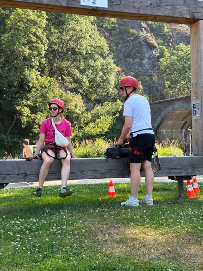 The image depicts two individuals wearing red helmets and safety harnesses, indicating they are likely at an adventure park or an outdoor activity center. The person on the left, a female, is sitting on a wooden beam and wearing a pink t-shirt and dark shorts. She has a white sling supporting her left arm. She is looking to her left and wearing sunglasses. The person on the right, a male, is standing and wearing a white t-shirt and black shorts. He is holding a black backpack. They are positioned in front of a wooded area with a rocky cliff face in the background. An arched stone bridge is visible in the distance. The ground is covered in green grass with scattered white flowers. Orange traffic cones are placed on the grass near the wooden beam. A smaller child is visible in the background, sitting on the beam to the left of the woman. There are QR codes and signage on a wooden post to the right of the standing man. The overall scene suggests a recreational outdoor environment. The location is identified as the city of Warisy, Belgium.