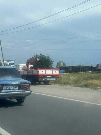 The image captures a daytime scene on a road in Ayntap, Armenia, viewed from inside a vehicle. In the immediate foreground, the rear of a light blue sedan, likely a Lada, is visible. Its Armenian license plate reads "34 LM 342". Behind this car, a red and white Mitsubishi truck with a crane attachment is stopped or moving slowly. The truck's rear displays the word "MITSUBISHI" vertically along its right side, and its license plate "04 FF 633" is partially visible.

The road itself is asphalt, with a white dividing line visible to the right. To the left of the road, there's a strip of green grass and some scattered trees, including a prominent green tree behind the Mitsubishi truck. In the background, under a bright, slightly hazy blue sky, several power lines stretch across the frame. Further in the distance, some industrial-looking buildings or structures can be seen, partially obscured by fencing and foliage. A billboard, partially visible on the left, shows the text "CHEVROLET". The weather appears clear and sunny.