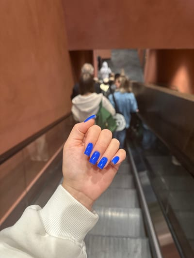 A first-person perspective shot shows a woman's hand with bright blue acrylic nails extended forward in front of an escalator. The woman is wearing a white sweatshirt with ribbed cuffs. In the background, the escalator is moving downwards, with several blurred figures visible on it, including a person in a light-colored hooded jacket and another in a denim jacket. The walls of the escalator shaft are a reddish-orange color. The location is not Yuzhnyye Koryaki, Russia.