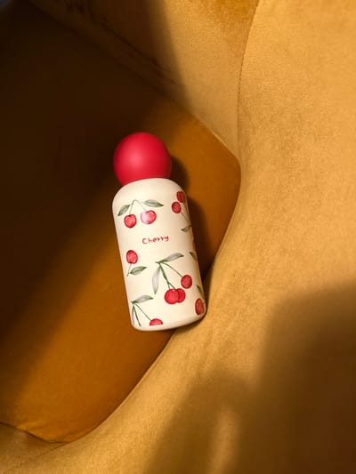 The image captures a white, cylindrical bottle with a red, spherical cap, resting within a mustard yellow upholstered environment. The bottle is decorated with multiple red cherry illustrations, including green stems and leaves, and the word "Cherry" is printed in red script on its surface. The surrounding material, likely fabric, displays folds and variations in shading, suggesting a soft, curved interior space. No individuals, actions, or interactions are present. The scene is situated in Wanneperveen, Netherlands.