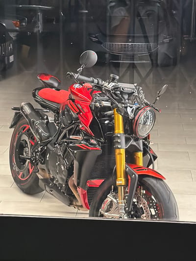 A red and black MV Agusta motorcycle is parked indoors, possibly in a showroom or garage. The motorcycle features gold-colored front forks, a circular headlight with a red accent ring, and black wheels with red detailing. An "Arrow" branded exhaust pipe is visible on the left side. Reflections in the background suggest it is a well-lit indoor space with other vehicles or structures present. The location in Novara, Italy, is not discernible from the image.