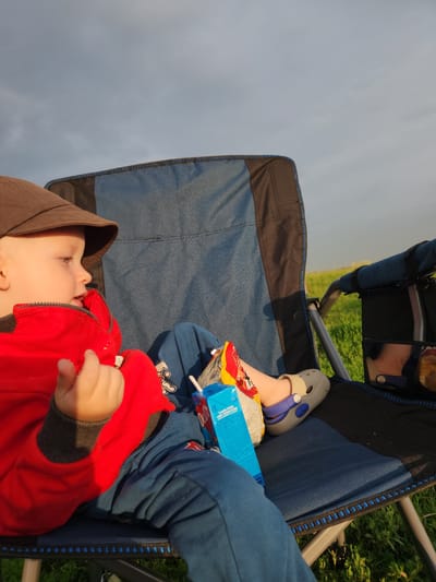 This media file captures a close-up, tilted view of a young child sitting in a blue and black camping chair. The child is wearing a red hooded jacket with grey trim, and a brown hood is pulled over their head. They are looking towards the left side of the frame, with their mouth slightly open, suggesting they might be talking or eating. One hand, resting on the jacket, is visible. The child's legs are bent, and they are wearing grey Crocs with blue accents, one of which is prominently visible. Beside the child's legs is a blue juice box with some visible text, and what appears to be a snack bag with red and yellow packaging.

In the background, out of focus, is a grassy area and a cloudy sky. The lighting suggests it is either early morning or late afternoon, casting a soft glow. The overall scene appears to be outdoors, likely in a park or natural setting, given the grass and camping chair. The composition focuses on the child, their immediate surroundings in the chair, and hints of the outdoor environment. The image conveys a sense of relaxed outdoor activity.