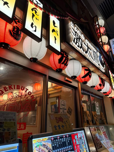 This is a nighttime, eye-level shot of a Japanese restaurant exterior, likely in Adachi, Japan. The scene is dominated by traditional Japanese lanterns, some glowing with orange light and others white with thin stripes. Several illuminated signs with Japanese text hang above, also contributing to the ambiance.
A prominent sign, featuring bold black lettering against a white background, reads "GEKIBUTOR" (though the OCR indicates "GEKIBUTOR" which is not Japanese, it could be a phonetic spelling). This sign appears to be a restaurant name. Below this, a vertical menu board is visible, displaying various food items and prices in Japanese. The text on the menu includes categories like "焼鳥" (yakitori), "冷菜" (appetizers), and "餃子" (gyoza). Other signs scattered throughout the image provide information like "喫煙OK" (smoking OK), "2階席あります" (second floor seating available), and business hours.
The overall impression is one of a lively, welcoming establishment, characteristic of many Japanese izakayas or restaurants. The warm glow of the lanterns and the detailed signage suggest it is a popular spot, likely busy with patrons enjoying a meal and drinks. The perspective and lighting create a slightly angled, dynamic view of the restaurant's facade.