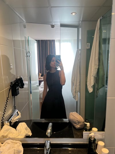 A female subject is depicted in a mirror selfie taken within a hotel bathroom. The image's reflection identifies the location as the Hotel de l'Abeille in Orléans, France. The subject, with dark, wavy, shoulder-length hair, wears a sleeveless black A-line dress with subtle vertical detailing and a thin bracelet on her left wrist. She holds a smartphone with a triple camera array in her right hand, taking the photograph, and has her lips pursed. The foreground shows a black vanity countertop with an integrated sink, dual chrome faucets, a wall-mounted black hairdryer with a coiled cord, white towels, a beige toiletry pouch, various pump-bottle toiletries, and two white disposable cups. Two towels (one light, one greenish-grey) hang on a hook or rail to the right. The background, reflected in the mirror, reveals a hotel bedroom with a bed featuring white linens, patterned brown and beige curtains, and a window allowing natural light. The bathroom ceiling features recessed lighting and a smoke detector.