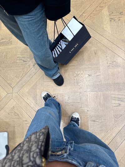 This is an overhead shot depicting two individuals standing on a light wood parquet floor within an indoor retail environment in Saint-Laurent-du-Var, France. The subject in the foreground wears light blue denim jeans and black and white low-top sneakers with "DIOR" branding visible on the sides. A segment of a brown and beige patterned item, consistent with Dior's Oblique motif, is partially visible at the bottom left. The subject in the background wears light blue denim jeans and plain black slip-on shoes. This individual holds a navy blue "DIOR" shopping bag, containing a white box and white tissue paper, by its black rope handles.