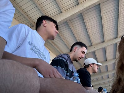 The image shows a low-angle perspective of three people standing under a roof, likely outdoors. A person in the foreground is wearing a white t-shirt with "Abercrombie and Fitch" on it. To their left is a person with a striped shirt, holding a water bottle, and next to them is a person with a white baseball cap and a black shirt. The background is dominated by the underside of the roof, made of repeating metal panels and support beams. There is no information to identify the specific location of this image in the city of Valpaços, Portugal.