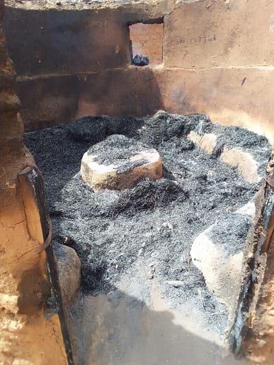 The image shows the interior of a small, rectangular room with mud brick walls and a dirt floor. The room is covered in black ash and debris, indicating that it has recently been burned. There is a small, rectangular window on the left wall, which is boarded up. A black plastic bag is visible through the window. In the center of the room, there is a large, round, rock-like object, partially covered in ash. The room is located at the coordinates 9.803 latitude and 8.646 longitude, which places it in the city of Kwal, Nigeria. There are no people present in the image.