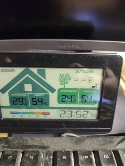 An indoor/outdoor thermometer and hygrometer is displayed on a black electronic device with a screen. The screen shows "INDOOR" and "OUTDOOR" readings. Indoor temperature is 29.2 degrees Celsius and humidity is 54%. Outdoor temperature is 24.2 degrees Celsius and humidity is 64%. The time displayed is 23:52. The device is situated on top of a dirty keyboard. The background appears to be a dark monitor or screen.