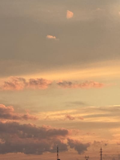 This image captures a sunset sky over Belokopitovo, Bulgaria. The sky is filled with clouds, some of which are tinged with pink and orange hues from the setting sun. The lower portion of the image shows a hazy horizon with visible power lines and pylons, suggesting an urban or semi-urban environment. There are no people or discernible actions depicted. The overall scene is one of a natural atmospheric phenomenon against a backdrop of human infrastructure.