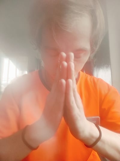 The image is a close-up, eye-level, medium shot of a person with fair skin and light brown hair, wearing an orange t-shirt and a thin silver chain necklace. The person's hands are pressed together in front of their face in a prayer-like gesture. The background is out of focus and appears to be indoors, with a bright light source visible on the left, suggesting a window. There is a slight haze or diffusion effect over the entire image. The exact location within the city of Salsk, Russia cannot be determined from the visual information provided.