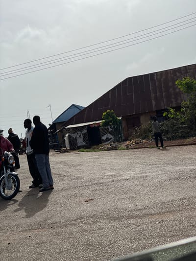 A group of individuals are gathered on a dusty, asphalt road in front of a dilapidated structure with a corrugated metal roof. The building appears to be a market stall or workshop, with various items scattered around its entrance, including debris and branches. To the left, a motorcycle is parked. One person is seated on the motorcycle, while others stand nearby, conversing. The sky is overcast, with faint clouds. The overall scene suggests a bustling, possibly informal, commercial area in Kassa Market, Nigeria.