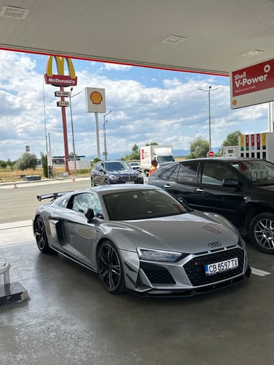 The image depicts a silver Audi R8 parked under a gas station canopy. To the left, a McDonald's sign is visible with a "McDrive" and "0-24" designation. A Shell V-Power sign is also present on the canopy. A dark-colored SUV is parked behind the Audi, and a white truck and other vehicles are in the background, suggesting a service station environment. The location is identified as a gas station in Drazhevo, Bulgaria. The ground is paved concrete, and the sky is partly cloudy.