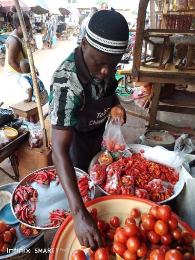 A man in a cap and apron labeled "Tobe Chinelafu" is selling produce at an outdoor market. He stands behind large bowls filled with red tomatoes and peppers. He holds a plastic bag and selects tomatoes to fill it. Other people are visible in the background, suggesting a busy marketplace setting. The bowls display the colorful produce prominently.