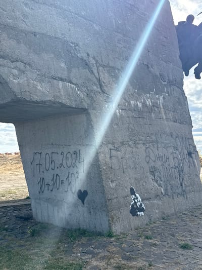 The image displays the lower section of a large, weathered grey concrete structure located in Izium, Ukraine. A strong, diagonal sunbeam cuts across the concrete from the upper right to the mid-left.
On the lower left face of the concrete, black spray-painted graffiti reads "17.05.2024" followed directly below by "10+10=10²." Beneath this text, a solid black heart symbol is spray-painted.
On the lower right face, additional black graffiti in Cyrillic script is present, with partially legible words including "Бро," "ДНИН БАЙДЯ," and "ТЕЛІНГГРОЙ." Below this text, a black and white stencil artwork depicts a crouched figure in a patterned robe.
The base of the concrete structure meets a paved surface of dark, irregular stones on the right, transitioning to dry earth with sparse green vegetation on the left. In the upper right background, a dark, indistinct sculptural element or figure is partially silhouetted against a partly cloudy sky.