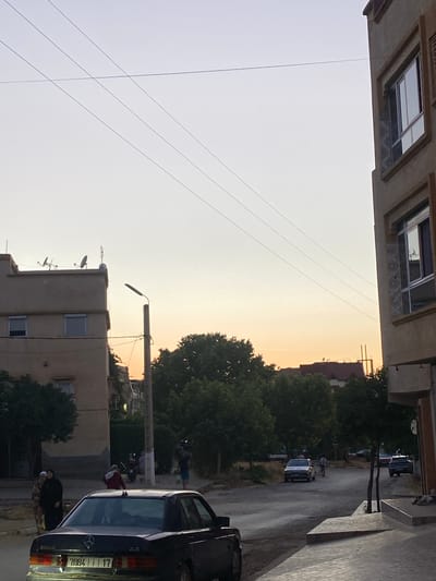 The image depicts a street scene in El Hajeb, Morocco, during what appears to be dusk. In the foreground, a dark-colored sedan, possibly a Mercedes-Benz, is parked. The vehicle has a license plate displaying the numbers "7884 17". To the left of the sedan, two individuals, one wearing a dark headscarf and black outer garment, and another in a patterned dress, are standing on what seems to be a sidewalk. Further down the street, several cars are visible, including a light-colored sedan and a darker SUV. A street lamp stands to the right of a building, casting a subtle light. In the background, a line of trees separates the street from residential buildings with visible windows. The sky is a pale gradient of light blue and orange, indicating the approaching twilight. Overhead, thin power lines stretch across the sky, supported by poles. The architecture of the buildings suggests a typical urban or suburban environment in Morocco.