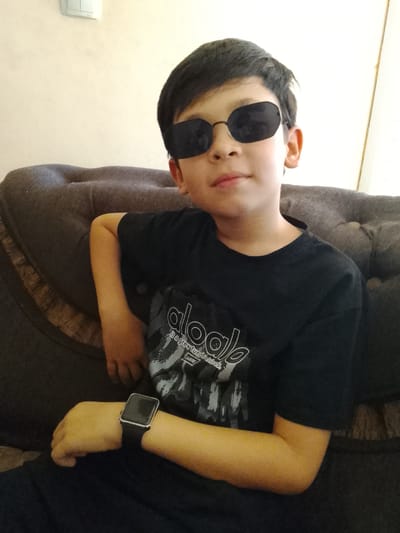 A young boy wearing black sunglasses and a black t-shirt with a white graphic print sits on a dark grey tufted sofa. He is looking directly at the camera, with his left arm resting on his leg. A smartwatch with a black band is visible on his left wrist. The background consists of a light-colored wall and a narrow window frame on the right. This scene is located in a room within Kasri Arifan, Uzbekistan.