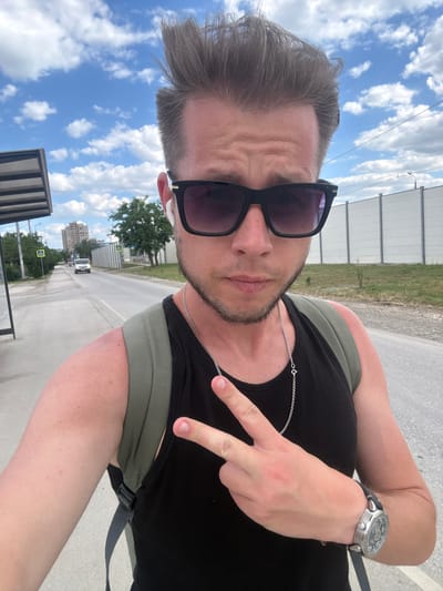 A man wearing sunglasses, a black tank top, a silver chain, and a backpack is standing on a paved area next to a road, outdoors. He is holding up two fingers in a peace sign. The background includes a street with a white van driving on it, a building, trees, a bus stop, and a white fence. The sky is blue with white clouds. This image was taken in Komsomolskoye, Ukraine.