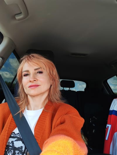 A woman with peach-colored hair, wearing an orange cardigan over a white t-shirt, sits in the driver's seat of a car. She is looking at the camera with a slight smile. Her makeup is visible, with pink lipstick and eyeshadow. A black seatbelt crosses her chest. The interior of the car is dark, with headrests of the back seats visible. The windows show a view of trees and a cloudy blue sky, suggesting it is daytime. To the right, a red, white, and blue jersey with the number "7" is visible, suggesting a sports context or affiliation. The scene appears to be in Alnashi, Russia.