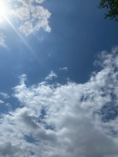 The image shows a bright blue sky with scattered white cumulus clouds. The sun is visible in the upper left corner, casting a bright glare. In the upper right corner, a small cluster of green leaves from a tree branch is visible, suggesting the photo was taken outdoors. The lower portion of the image is dominated by larger, more textured clouds, some appearing darker and heavier. The overall scene depicts a daytime sky with typical cloud formations. The provided context places this scene in the city of Важное, Russia.