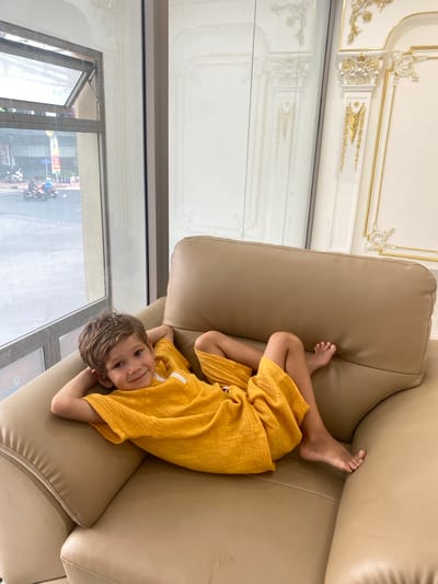 A young boy with light brown, wavy hair smiles at the camera while lying on a tan leather couch. He is wearing a yellow, textured t-shirt and yellow shorts. His left arm is bent behind his head, and his legs are extended. Through a large window to the left of the couch, a street scene in Phú Thủy, Vietnam, is visible, with motorcycles and buildings in the background. The wall behind the couch features ornate gold and white decorative molding.