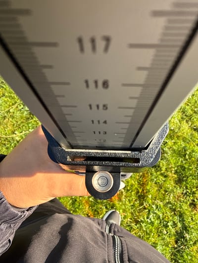 The image shows a close-up of a leveling staff held by a person's hand, with a leveling bubble visible at the bottom of the staff. The staff has numerical markings, with "117", "116", "115", "114", "113", and "112" clearly visible. The background consists of green grass, indicating an outdoor environment. The image is cropped to focus on the measuring instrument and the hand holding it. The location is specified as Yundola, Bulgaria.