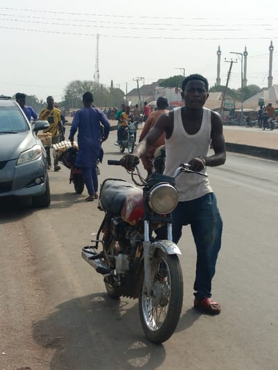 A man in a white tank top and jeans stands next to a motorcycle on a road. Other people are visible in the background, including a vendor carrying trays of packaged goods. A gray car is parked to the left. In the distance, minarets rise, and a cell tower is seen behind a tree. The scene takes place during the day, with bright light casting shadows on the road.