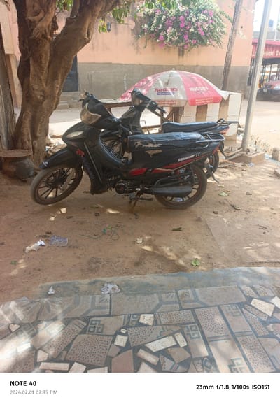 The image depicts an outdoor scene in Tudun Wazurchi, Nigeria, captured during daytime on 2026.02.01 at 02:35 PM. The foreground features a tiled ground surface composed of irregular, fragmented tiles in patterns of beige, brown, and blue-green hues. Above this, a dirt and sandy ground area extends into the midground, scattered with debris including plastic wrappers, paper, and dry leaves.

Two motorcycles are parked centrally. The prominent motorcycle in the foreground is black with red accents, featuring "WAPED" and "VIBRANT II" branding on its side and "CAS220-60" on the engine cover. Its seat is covered with a worn black material, and it rests on a kickstand. A second, dark-colored motorcycle is partially visible behind it.

To the left, a large tree trunk with exposed roots anchors the scene, and an earthenware pot rests at its base. In the mid-background, a light orange-peach colored building wall is visible, with a dark doorway partially obscured by the tree. Above the wall, flowering bougainvillea with pink and white blossoms cascades. To the right, a red and white "HUGGIES" branded umbrella is open, positioned over what appears to be a table constructed from concrete blocks. A utility pole stands on the far right. The background on the right reveals a street or pathway with glimpses of other structures and a vehicle. No individuals are present, and no actions or interactions are depicted.