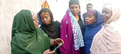 The image captures six individuals, five adult females and one adult male, gathered outdoors in Dan Maliki, Nigeria. In the foreground, an adult female on the left, wearing a dark green patterned hijab and eyeglasses, holds a light green smartphone in her left hand, displaying its screen towards the group. Her right hand, visible below the phone, exhibits distinct henna patterns. She appears to be addressing the others. To her right, a second adult female, wearing a black hijab over a patterned head covering, observes the smartphone screen. Adjacent to her, a third adult female in a vibrant purple patterned garment and dark head covering is visible, wearing a gold-colored necklace. Next, a fourth adult female in a blue head covering and a patterned headband has her mouth open, indicative of speech or a reaction. On the far right, a fifth adult female in a beige patterned hijab smiles while looking towards the center of the group's interaction. Partially obscured in the background behind the third and fourth women is an adult male wearing a dark shirt. The setting features a light-colored, textured wall in the background.