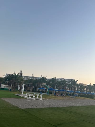This media file shows a view of a resort or hotel area with a lawn, palm trees, and buildings. The scene is dominated by a clear, pale blue sky that transitions to a hazy yellow or white near the horizon, suggesting either dawn or dusk. In the mid-ground, there is a row of buildings with white facades and visible windows. In front of the buildings, a line of blue umbrellas and lounge chairs are set up on a gravel or sand path. Several palm trees are scattered throughout this area, casting shadows. The foreground features a strip of green grass. There appear to be a few people in the lounge chairs, some of whom are wearing swimwear, suggesting a recreational setting like a beach resort or a pool area. The overall atmosphere is calm and relaxed. The image is oriented vertically. No visible text is present. The location is identified as Rhodes, Greece.