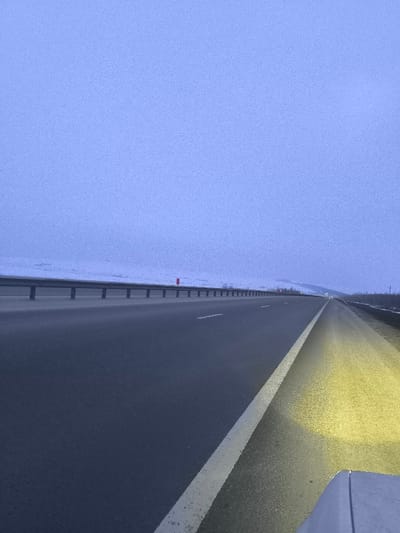 The scene depicts an empty multi-lane asphalt highway under a pale blue-grey, overcast sky, likely at dusk or dawn, in the vicinity of Pobeda, Russia. The viewpoint is from the front right of a vehicle, with a portion of its hood or bumper visible in the lower right quadrant, casting a bright yellow light beam onto the immediate right shoulder. The highway extends into the distance, flanked on the left by a metallic guardrail separating it from a snow-covered landscape of low hills or fields. A single red marker post is visible on the snowy embankment beyond the guardrail. The road surface is clear, marked with white dashed lane lines and a solid white line defining the right shoulder. No individuals or other vehicles are visible within the frame.