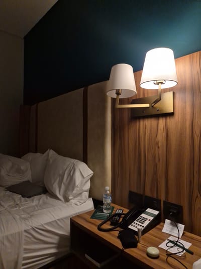 The scene is a hotel room in Veracruz, Mexico. The room features a bed with white linens and pillows. Next to the bed is a wooden nightstand. On the nightstand, there is a black telephone, a bottle of water, some papers, a pen, and a small, round, white object. Mounted on the wall above the nightstand is a wooden panel with a brass double-arm lamp. The wall is divided into two sections: one is a dark teal color, and the other is a light beige color.