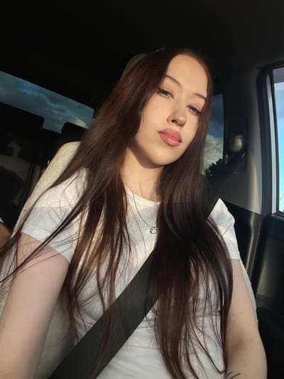 A young woman with long, dark brown hair is in the passenger seat of a car, taking a selfie. She is wearing a white t-shirt and a black seatbelt. The sun is shining, casting a warm glow on her face, and the sky is visible through the car window. Her eyes are closed, and her lips are slightly parted, suggesting a moment of relaxation or a playful pose. The scene appears to be set during the daytime, likely late afternoon due to the golden hour lighting. The background shows the interior of the car and a glimpse of the sky and trees outside, indicating she is likely on a road trip or a scenic drive. The overall mood of the image is relaxed and casual. No visible text is present in the image. The location context of Gavrilovka, Russia, does not provide specific visual cues within the image itself.