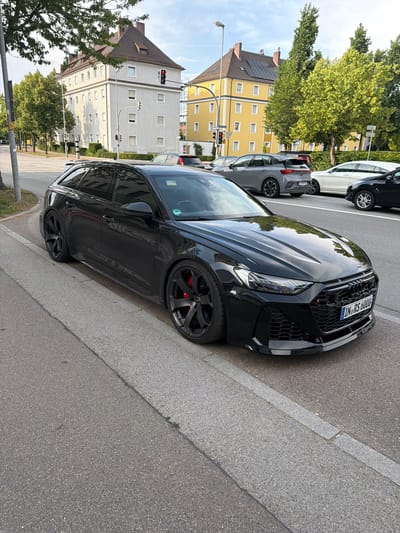 A black Audi RS6 sedan is parked on the side of a street in Ingolstadt, Germany. The car is facing towards the right side of the frame, with its front end prominently displayed. It has a sleek, sporty design with a black grille and aggressive front bumper. The wheels are black, and red brake calipers are visible behind the spokes.
In the background, there are several multi-story apartment buildings with light-colored facades and numerous windows. Some of the buildings have red-tiled roofs. Trees with lush green foliage line the street and the sidewalk in front of the buildings. A traffic light is visible, showing a red signal. The sky is partly cloudy.
The scene appears to be set during the daytime. The overall impression is a typical urban street scene in a European city. The car is the central focus, suggesting it might be the subject of interest, perhaps due to its performance or luxury attributes. There is no discernible activity or event taking place other than the car being parked. No people are visible in the frame.
Visible text on the car's license plate reads "IN-RS 6000". There are also some small stickers on the windshield, but the text is illegible. Other signs in the background are also too small to read. The weather appears to be mild and pleasant, suitable for outdoor activities. The mood is neutral, reflecting a normal street scene.
