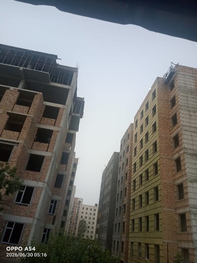 The image shows a construction site in Bokhtar, Tajikistan, with several multi-story buildings under various stages of completion. In the foreground on the left, a concrete and brick building is under construction, with exposed rebar at the top. Its exterior walls have a pattern of brickwork interspersed with concrete frames for windows and balconies. To the right, two more buildings are visible. One is clad in yellow insulation material and brick, with rows of dark window openings. The other building to the far right is also under construction, with concrete framing and some brickwork. Further in the background, a completed residential building with multiple windows is visible. The sky is overcast, indicating a cloudy or hazy day. The timestamp at the bottom left corner reads "2026/06/30 05:16," identifying the photo as taken on June 30, 2026, at 5:16 AM.