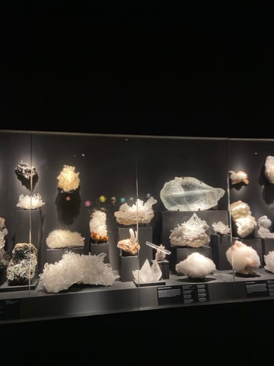 A display case in a dimly lit room, containing various mineral specimens, including several clear quartz crystals. The display is lit from above, and the specimens are on dark, rectangular pedestals, and are arranged in a grid-like pattern. The specimens are mostly white, but there is one specimen in the bottom left corner that is a dark grey. There are three small, multi-colored specks in the middle of the case. There is also a small label at the bottom of the case on the right. The location is a museum in Greenwich, Connecticut. There are no people in the image.