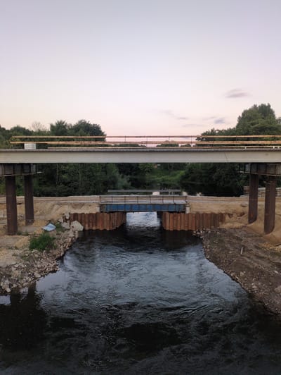 The image shows a construction site for a new bridge over a river in Посёлок Молочной фермы, Russia. The river is dark and flowing, with ripples and some small whitecaps. The riverbanks are lined with rocks and dirt, with some vegetation growing on them.
There are two bridges visible in the image. The lower bridge is an older, blue bridge that appears to be in use, though it is partially obstructed by construction materials. The upper bridge is a new, unfinished bridge, likely for a highway, made of concrete and steel. It is supported by large, brown metal pillars. The bridge is undergoing construction, with wooden scaffolding and construction materials visible. The surrounding area is a mix of dirt, sand, and some gravel.
The sky is a pale pink and blue, suggesting either dawn or dusk. Trees and foliage line the background, with more trees visible on the opposite bank of the river. There are no people or vehicles visible in the image. The overall scene depicts an active construction zone where infrastructure is being developed.