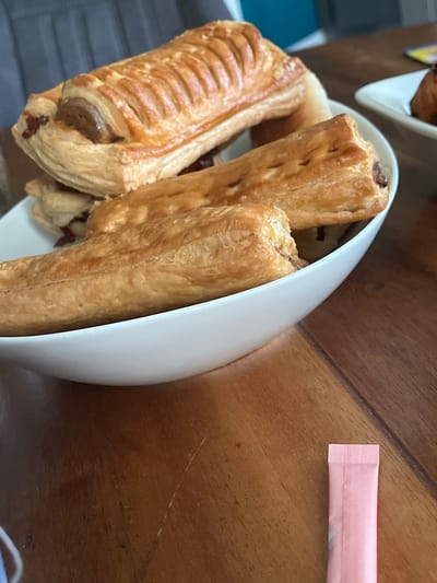 The image shows three pastries in a white bowl, placed on a wooden table. The pastries appear to be savory, with a flaky golden-brown crust and a filling that seems to be sausage or meat. One pastry in the foreground is partially visible, showing its ribbed crust and the dark filling peeking out. Behind it, two more pastries are arranged, one slightly to the left and the other to the right. The bowl is white and has a curved edge. To the right of the bowl, a pink packet, possibly containing sugar or sweetener, is visible on the table. The wooden table has a visible grain pattern and some light scratches. The overall setting seems to be indoors, likely a casual meal or snack. There are no people visible in the image. The lighting suggests it could be daytime, but no specific time of day can be determined. The scene is set in Uithuizen, Netherlands, but there are no specific visual cues to confirm this location within the image itself.