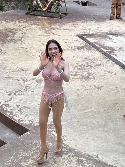 A young Asian woman is posed mid-dance on a textured concrete surface, which appears to be an outdoor performance area in Nong Prue, Thailand. She wears a sparkling pink two-piece outfit with fringe detailing and high-heeled sandals. Her hands are raised in a welcoming gesture, and she has a wide smile. The background shows a decorative structure with lights and foliage, a large bucket, and a portion of another person's legs clad in elephant-patterned trousers. The ground is a mix of light-colored concrete with areas of discoloration and cracks, suggesting it is well-used.