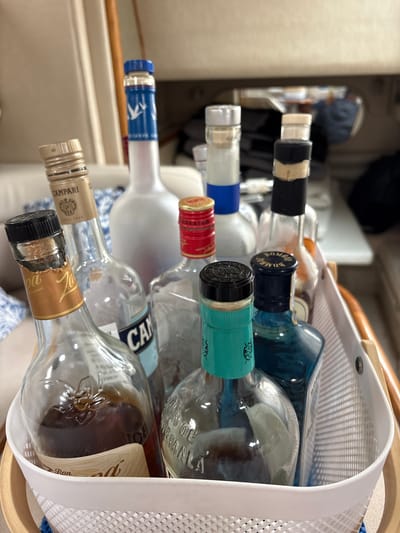 A shallow depth of field, close-up shot captures a white wire basket filled with several empty and partially filled liquor bottles. The bottles are of various shapes and sizes, with labels indicating brands like Campari, Ron Zafra, and what appears to be Bombay Sapphire. One bottle is a clear glass with a brown liquid, possibly rum, and an embossed floral design. Another bottle has a light blue liquid with a teal-colored label. Behind these are larger, mostly empty bottles, including a clear bottle with a blue label that reads "CAMPARI" and a tall, frosted white bottle with a blue cap and a logo featuring a goose. The background is blurred, suggesting an indoor setting with neutral-toned furniture and possibly a window. The overall impression is one of a collection of used liquor bottles, possibly awaiting disposal or recycling, in a domestic or hospitality environment. The location in La Punta, Peru, is not discernible from the visual information in the image.