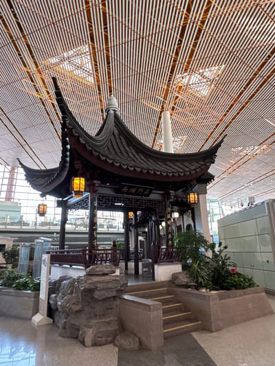 The image shows a traditional Chinese structure indoors, possibly a pavilion or decorative element, within a modern building, identifiable as the Beijing Capital International Airport in Shunyi District, China. The structure has a curved, tiled roof with intricate detailing, lit by traditional lanterns. The building features a latticed ceiling. In the background, glass walls reveal views of the modern airport. To the right of the Chinese structure is a stepped platform with vegetation, and a rock feature with signage.