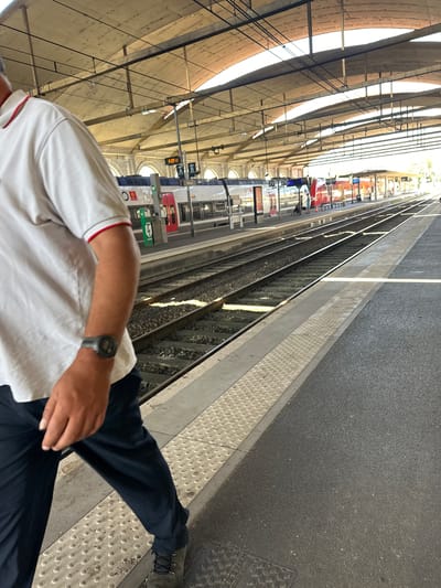 The image captures a portion of a train station platform in Nîmes, France, under a covered, arched ceiling. In the foreground, a person wearing a white polo shirt with red trim and dark pants walks along the platform, their left arm visible, showing a dark digital wristwatch. The platform features a textured, grey surface with a tactile warning strip along the edge, separating it from the train tracks. The tracks are visible, with gravel ballast and sunlight glinting off the rails. In the mid-ground, several train cars are stopped at the platform, displaying red and white livery. One train car has an electronic display showing "6:09 13" and destination indicators "A B" and "Nîmes". The station appears to be an open-air structure, with sunlight filtering through the gaps in the roof. The overall scene depicts a typical operational moment at a train station.