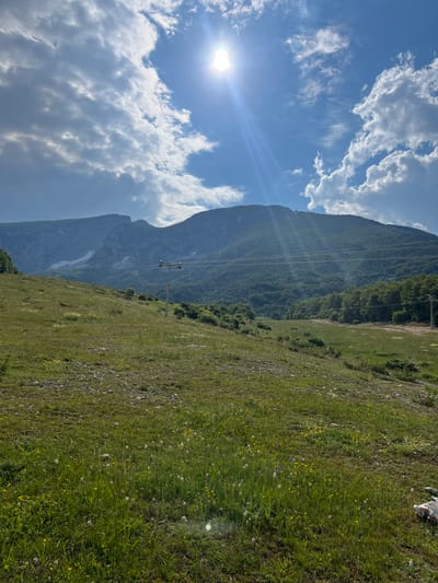 The image depicts a mountainous landscape under a bright, partly cloudy sky. The sun is visible on the left, casting a lens flare and illuminating the clouds. The scene is characterized by a steep, grassy slope dotted with small wildflowers, transitioning into a rocky area on the left. In the background, a large, imposing mountain dominates the view, with a patch of snow visible on its side. A line of power lines stretches across the mountain face, supported by utility poles. The overall impression is one of a serene, natural environment, likely during the daytime, with fair weather. There is no visible text in the image.