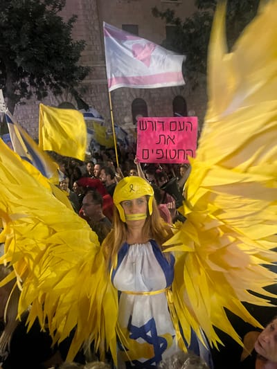A woman stands centered in the foreground, wearing a costume featuring large yellow feathered wings, a yellow helmet adorned with a yellow ribbon, and a yellow tape strip horizontally covering her mouth. Her long, light brown hair is visible beneath the helmet. She wears a white and blue dress with a prominent blue Star of David (Magen David) on the skirt and a yellow sash around her waist. She is part of a dense crowd, visible behind and around her, consisting primarily of adults. Among the crowd, various flags are displayed, including multiple Israeli flags (blue and white with a Magen David), numerous plain yellow flags, and a white flag with a pink inverted triangle held aloft on a pole. A pink protest sign, elevated in the background, displays Hebrew text: "העם דורש את החטופים" (The people demand the hostages). The scene is set at night, illuminated by artificial lighting, against a backdrop of a stone building featuring arched windows, characteristic of traditional architecture in the area. This event is occurring in the city of West Jerusalem, Israel, during what appears to be a public demonstration.