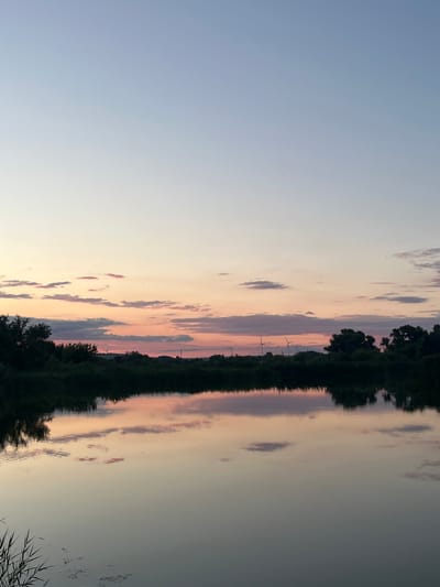 The image depicts a tranquil scene at sunset over a body of water, likely a river or lake, in Kotovo, Russia. The sky is a gradient of pale blue at the top, transitioning through soft pinks and oranges where scattered clouds are visible, reflecting in the calm water below. A dark silhouette of trees and foliage lines the far bank of the water, with several wind turbines visible in the distance on the horizon. The water's surface is smooth, mirroring the sky and the distant landscape. There are no people or animals visible in the frame, and the overall impression is one of peacefulness and natural beauty.