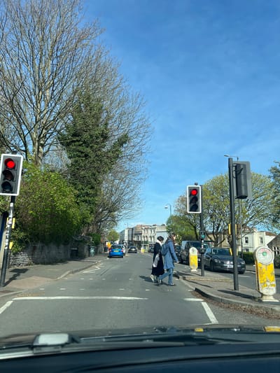 The image shows a street scene in Bristol, United Kingdom. Two pedestrians are crossing a street at a crosswalk, one appears to be a child/teenager. A traffic light is red, indicating that the pedestrians have the right of way. Cars are stopped at the intersection. Trees line the street, some bare and some with green foliage. Buildings are visible in the background. The sky is blue with minimal cloud cover.