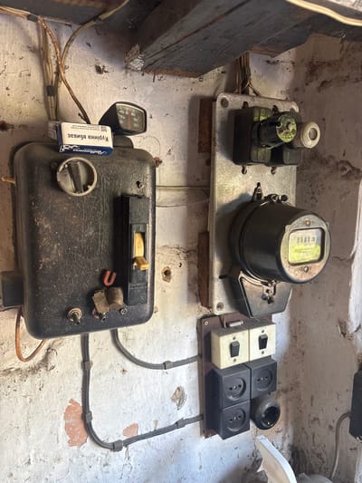 The image depicts an indoor scene, possibly a utility room or basement, in Pereiaslav, Ukraine. The subject is an old, wall-mounted electrical panel. Dominating the frame is a large, black, rectangular box with a main circuit breaker lever and various other electrical components. Attached to the wall to the right is a metal plate holding a fuse holder, a black cylindrical object that appears to be an electricity meter with a glowing green display, and below it, a block with two toggle switches and four electrical outlets. The walls are of rough, white-washed brick, showing signs of age and wear. Wooden beams are visible at the top of the frame. Electrical wiring is neatly but visibly routed along the wall. The overall impression is one of an aged, functional electrical installation. No people are present.